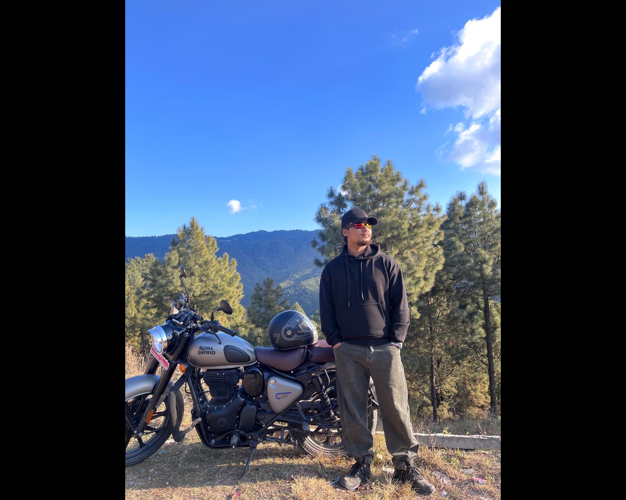 Rider standing beside a silver Classic 350 parked on a ridge, tall pines and blue hills behind