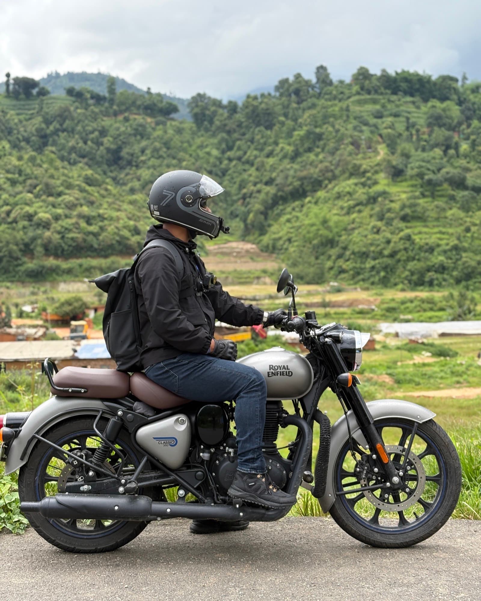 Rider in black gear sitting on a silver Classic 350 at the roadside, terraced green hills behind under a grey monsoon sky