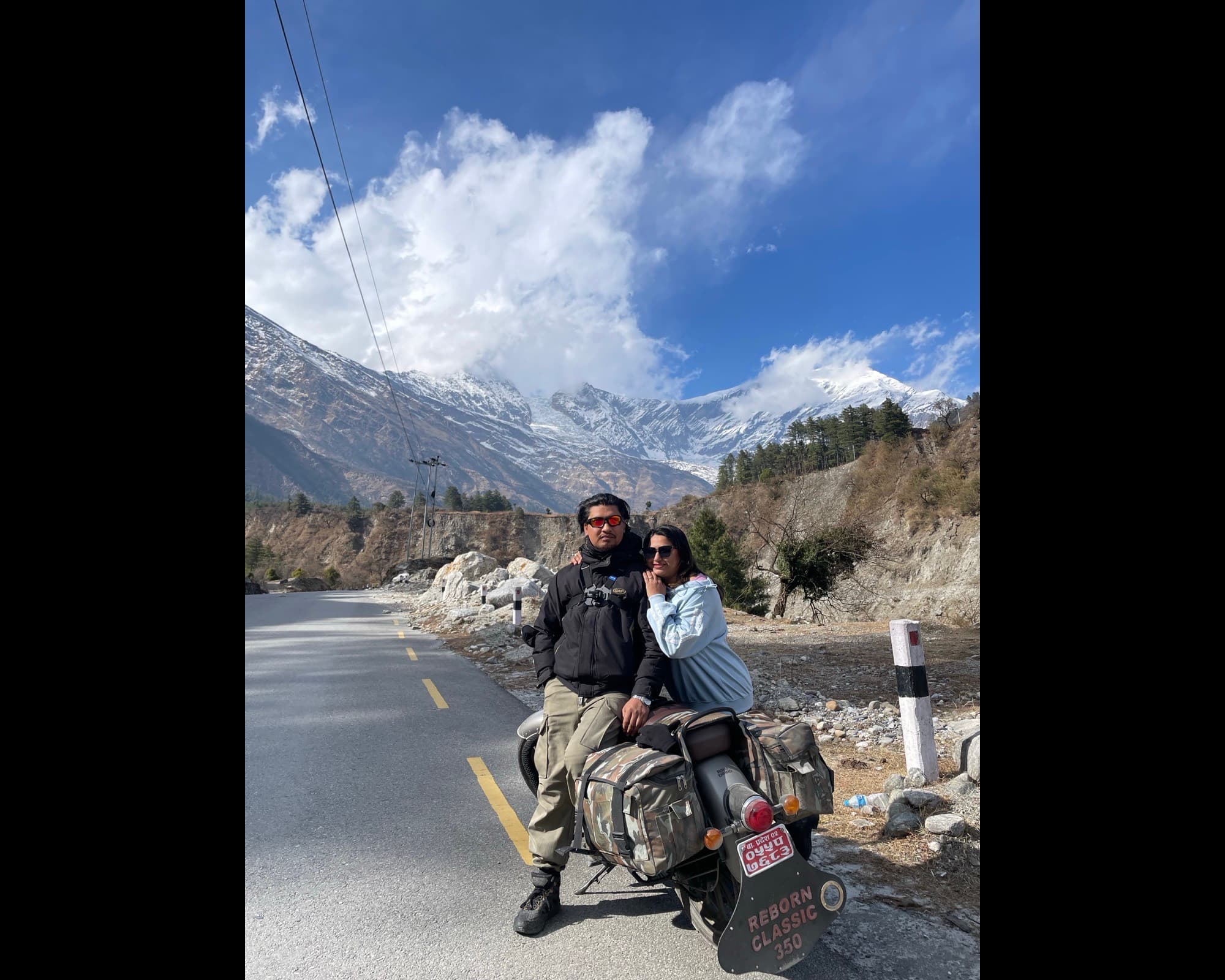Two riders sitting on a loaded Classic 350 on an empty mountain road, snow peaks rising behind