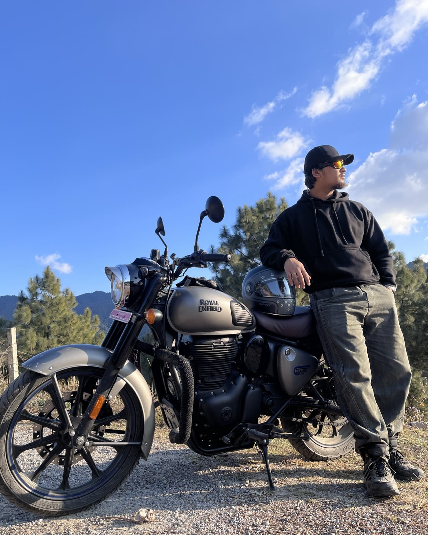 Ranjit in a black hoodie, cap and sunglasses, leaning against his gun grey Royal Enfield Classic 350 on a gravel pull-off, helmet resting on the seat and pine-covered hills behind